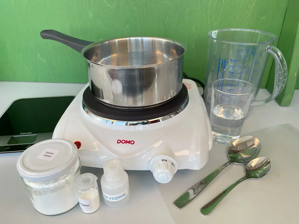 Against a green background on the left are kitchen scales, next to them a saucepan on a single hob, in front of it a screw-top jar with cornflour, a small glass container with gelatine powder, a small plastic bottle with glycerine, two spoons, and on the right in the background a glass of water and a measuring jug.
