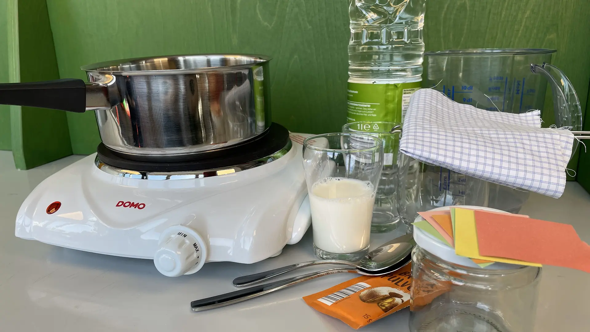 Glasses, a glass of cow's milk, a pot, a few spoons, a funnel, a hotplate, a measuring cup, a piece of cloth, a glass, vinegar, baking powder, scraps of paper and a jam jar.