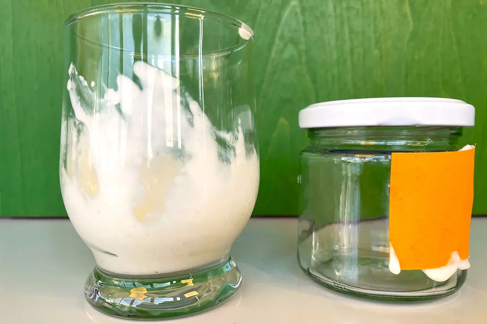 On the left is a glass with a white creamy substance next to a smaller glass jar with a white screw-on lid and an orange piece of paper stuck to it. The glasses stand in front of a green background.