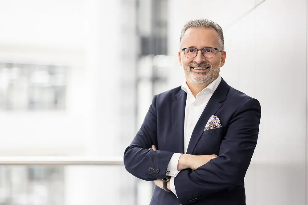 Carsten Knobel wearing a business suit and standing with arms crossed in a bright, modern office environment.