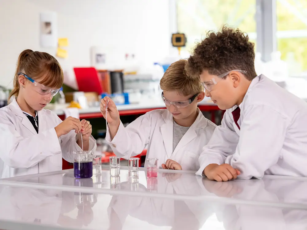Three children wearing lab coats and safety glasses conduct an experiment with colored liquids.